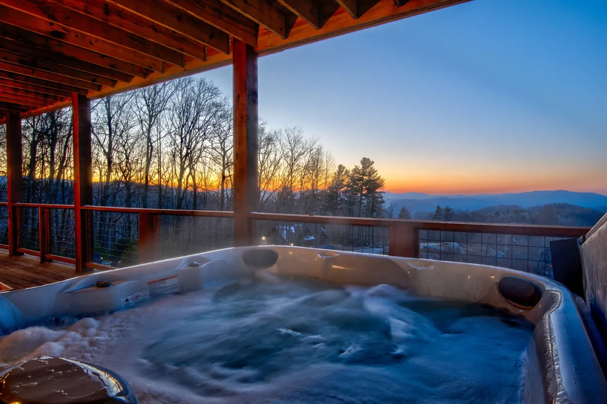 This exterior image centers on a hot tub perched on a deck, with sweeping mountain views and a vibrant sunset in the background. The tranquil ambiance is perfect for relaxation, and the setup invites stargazing or romantic evenings outdoors.