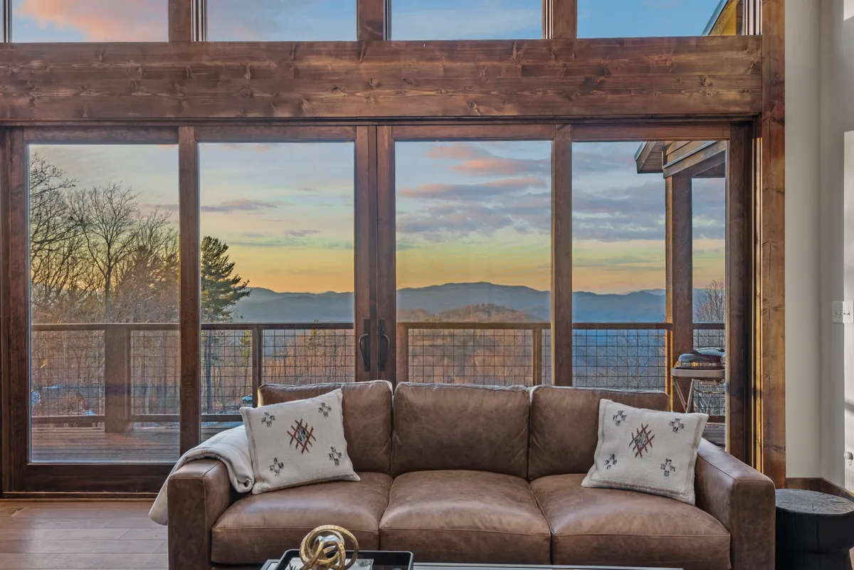 A cozy seating area with a leather sofa and decorative pillows positioned beside floor-to-ceiling glass windows, offering uninterrupted views of the mountains. The space is designed for relaxation, reading, or simply soaking in the tranquil beauty outside, merging comfort and scenery.
