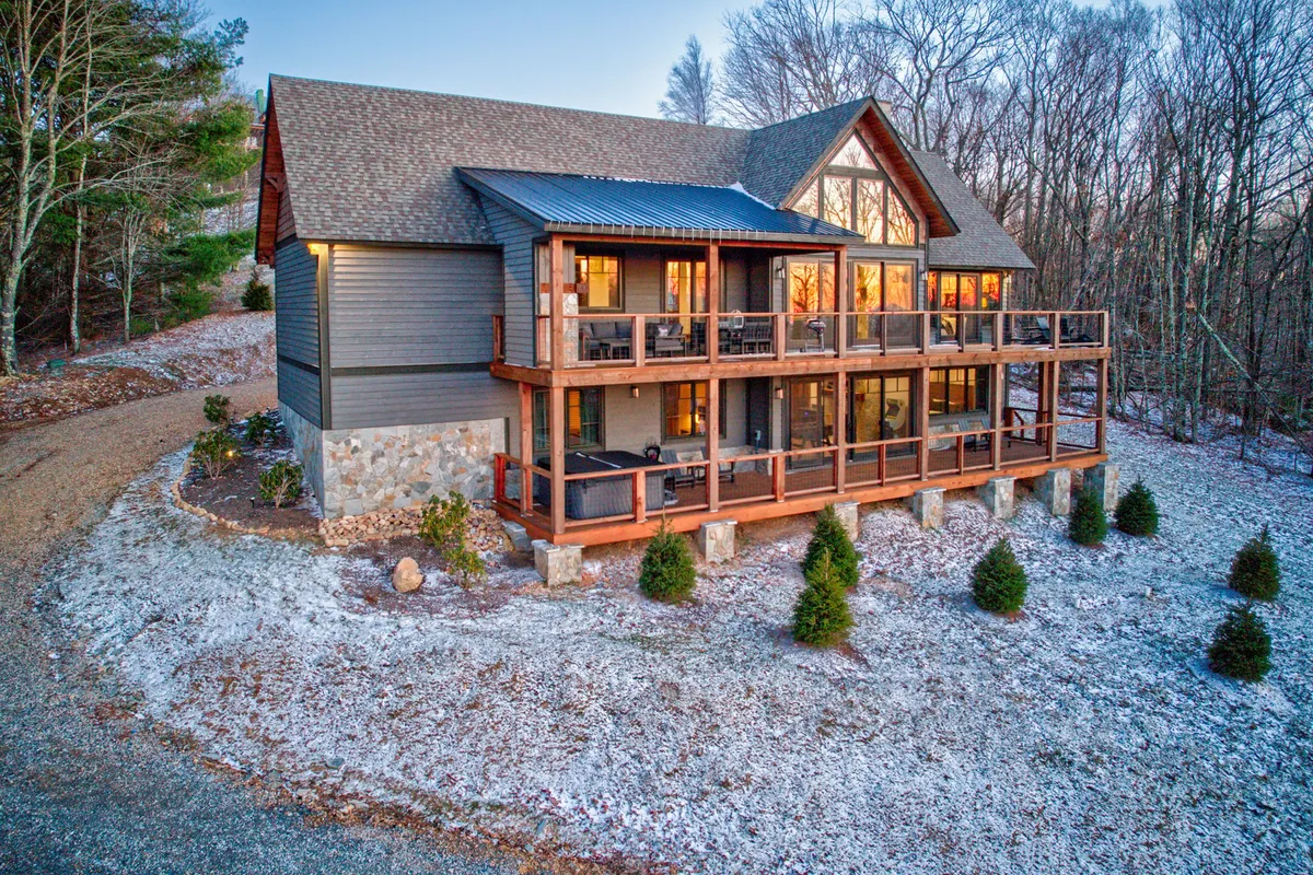 A welcoming exterior shot of the cabin, featuring a wooden balcony, large windows, a sturdy stone foundation, and lush greenery surrounding the property. The inviting balcony overlooks scenic views, suggesting a perfect spot for family relaxation or group gatherings after a day of adventure.