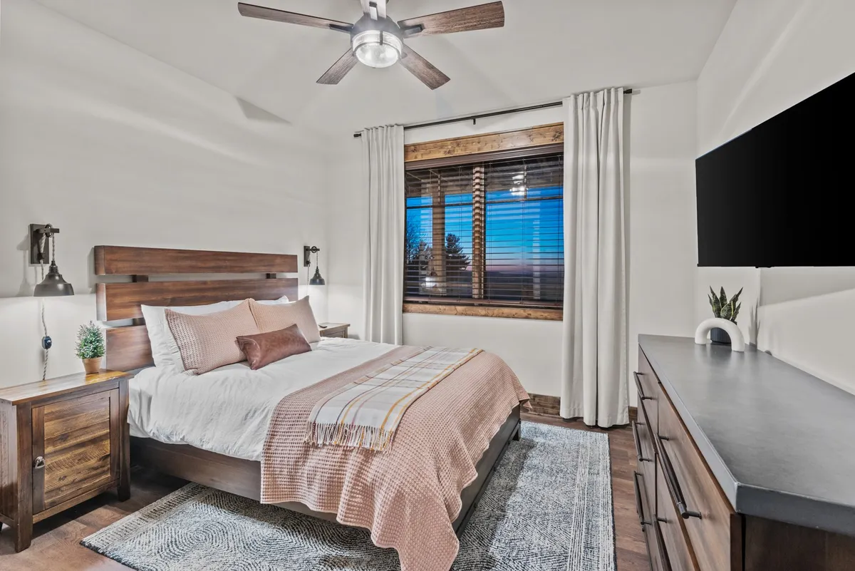 A serene bedroom featuring a large bed with a decorative throw, a spacious window with blinds allowing plenty of natural light, a wooden nightstand with a plant, and wall-mounted lamps. The tranquil setting suggests comfort and privacy, ideal for restful evenings.