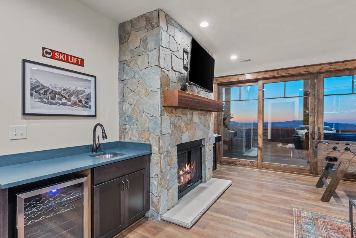 A warm and inviting living room with a stone fireplace as the focal point, large windows framing breathtaking mountain views, and a ski lift sign adding a playful mountain lodge touch. Plush seating creates an inviting atmosphere perfect for relaxing or entertaining, and the sunset glow through the windows hints at the seamless indoor-outdoor connection.