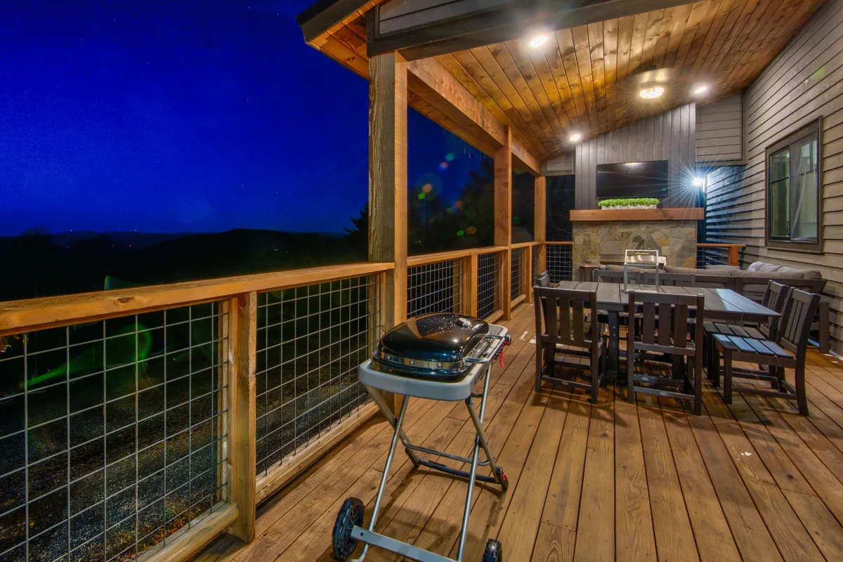 An outdoor deck scene with a grill, dining table, and an outdoor fireplace, providing an inviting setup for barbecuing, alfresco meals, and socializing under the stars. The wooden deck and ambient lighting create a festive, communal atmosphere.