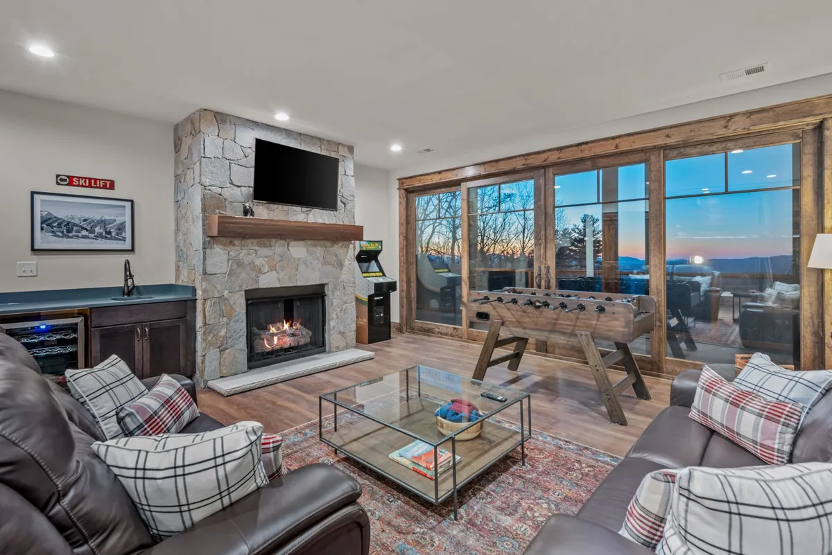 A warm and welcoming den featuring a stone fireplace, large windows that let in natural light, a game table, and a plaid sofa. The room exudes comfort and is set up for both relaxing by the fire and enjoying games with friends or family.