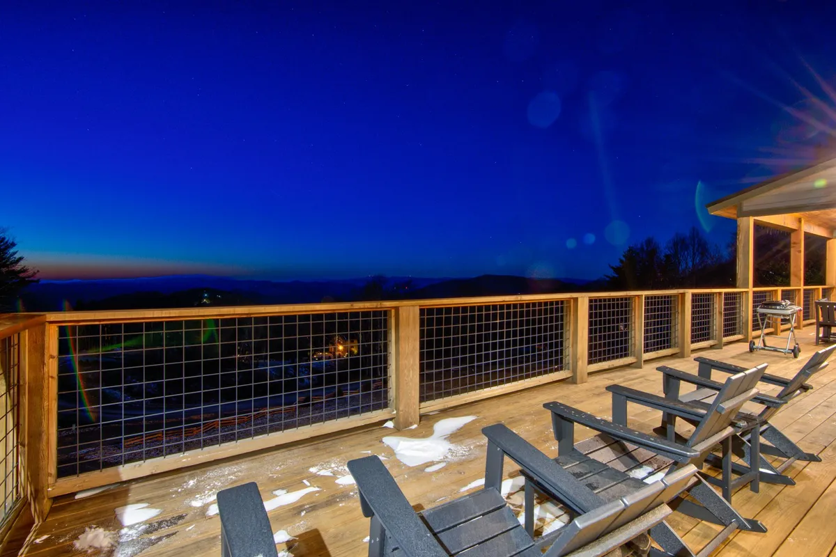 A tranquil nighttime scene featuring a wooden deck, Adirondack chairs, and a view of the star-filled sky. The setup is perfect for stargazing, evening relaxation, or sharing warm drinks by the fire pit. The outdoor environment suggests a connection to nature and the mountain atmosphere, resonating with experiences unique to each season.