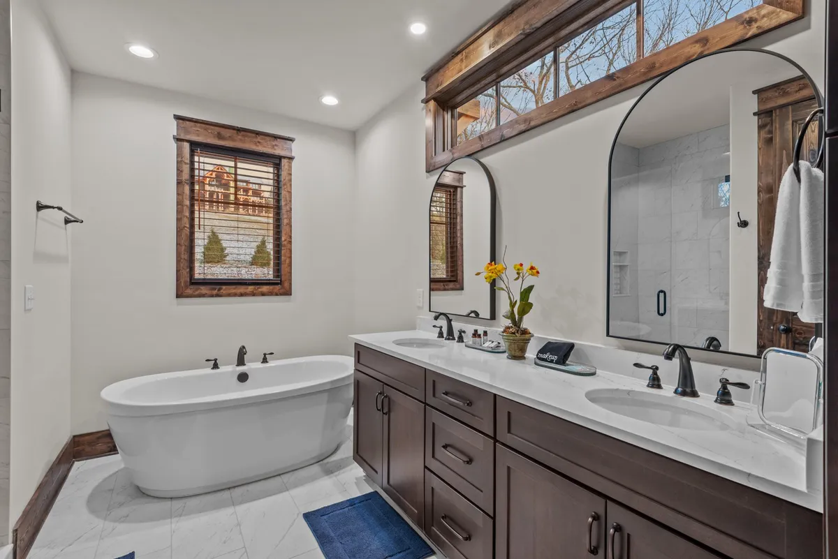 A luxurious en-suite bathroom featuring a freestanding soaking tub, double sink granite vanity, large mirrors, and a window with a scenic view. The space is bright, elegant, and spa-inspired, perfect for unwinding after an active day.