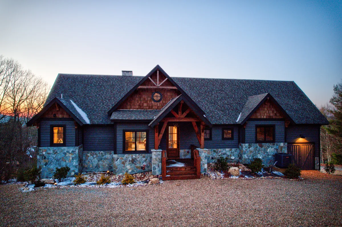 This exterior image showcases the lodge’s striking stonework, warm wooden beams, and large windows glowing with inviting light. The porch area, ideal for gatherings, overlooks a scenic outdoor setting that hints at mountain views and seasonal changes. The warm lighting suggests relaxation and social activity both inside and outside.