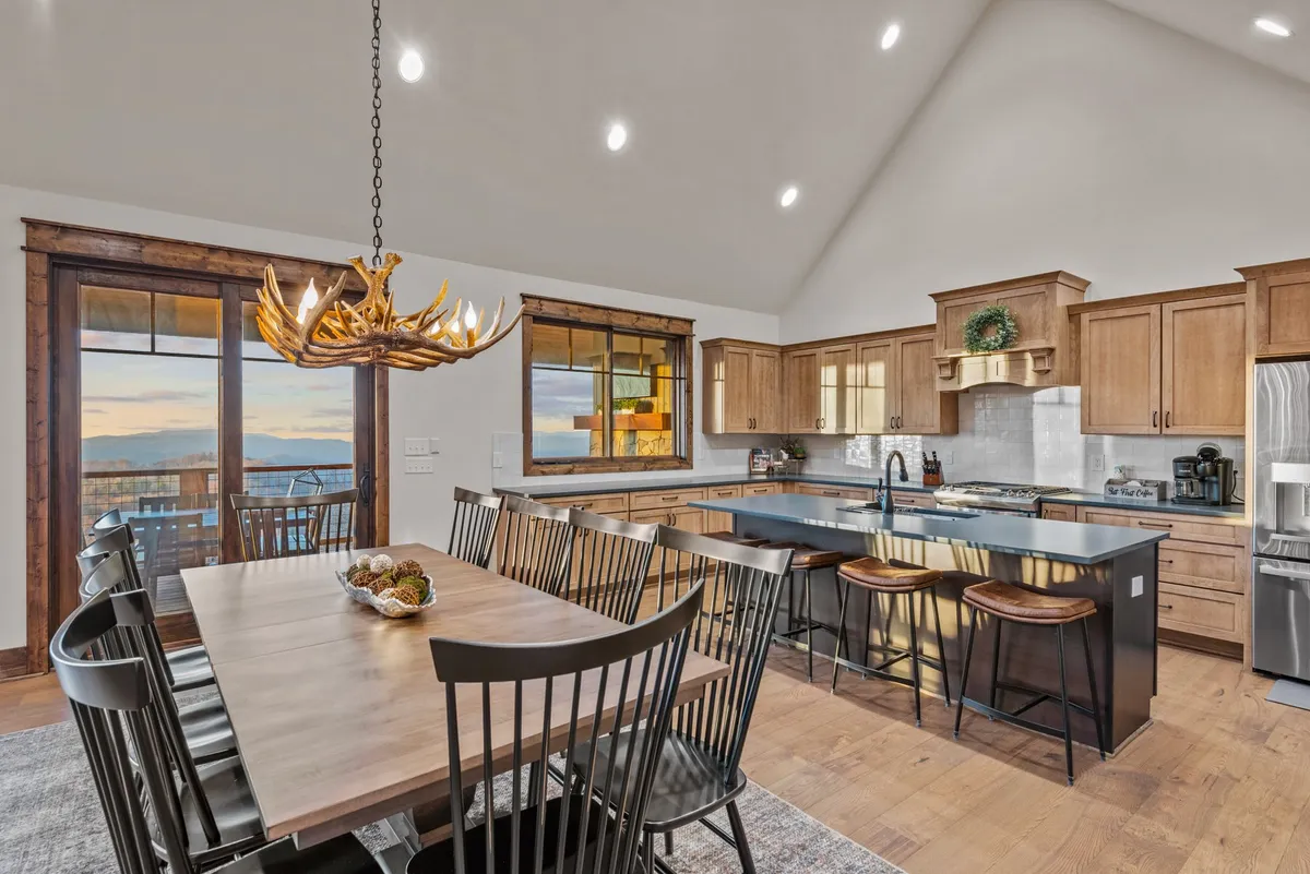 A spacious open-concept area combining a large dining table, a kitchen island with bar stools, and panoramic windows that capture the surrounding mountain landscape. The setting is ideal for family meals, cooking together, and enjoying laughter-filled gatherings bathed in natural light.