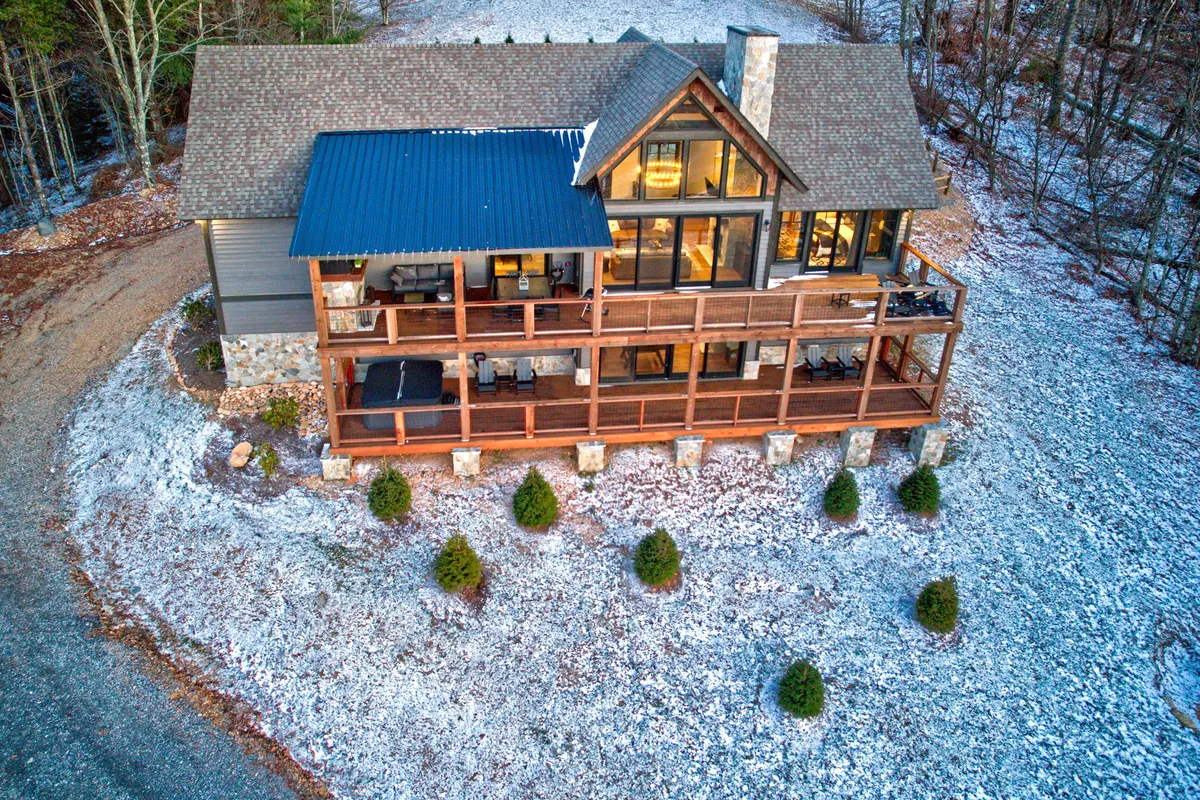 A wide wooden upper deck with large glass windows and a stone chimney, looking out onto a snow-dusted landscape. The deck is spacious and inviting, ideal for group gatherings, stargazing, or enjoying fresh mountain air as the day turns to evening.