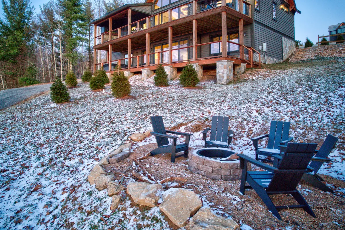 This exterior scene showcases a welcoming fire pit surrounded by Adirondack chairs on a deck. The arrangement encourages gathering and conversation, with the house subtly visible and trees framing the space, suggesting evenings spent fireside under the stars.
